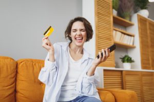 Young woman using mobile phone for online shopping, holding credit card and celebrating never paying full price